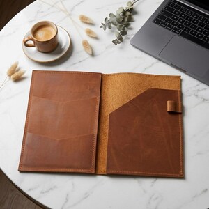 May include: A tan leather notebook cover featuring an open design with multiple interior pockets on the left side and a single pocket with an attached pen loop on the right side, showcasing visible stitching along the edges of the leather.