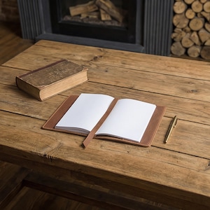 May include: An open notebook with blank white pages and a brown leather cover lies on a wooden table, next to a vintage book and a gold pen. The notebook has a brown bookmark. The table has a rustic, aged appearance.