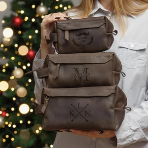 May include: Three brown leather wash bags stacked. The top bag features a mustache graphic and the text "BEST DAD EVER". The middle bag is engraved with "MORGAN". The bottom bag has the initials "LKN" with an arrow design.
