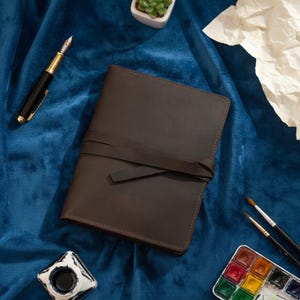 May include: A brown leather journal with a wrap-around tie closure. A gold and black fountain pen, inkwell, watercolor palette, and paintbrushes are arranged on a blue velvet surface. A small succulent plant in a white pot and a piece of crumpled paper are also present.