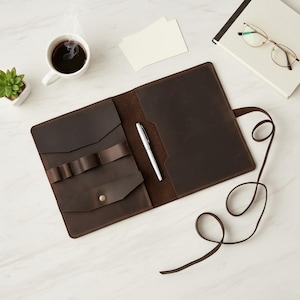 May include: Open brown leather portfolio with a pen, small cards, and a notebook with glasses. The portfolio has multiple pockets and pen holders. A cup of coffee and a small succulent plant are also on the white marble surface.