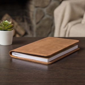May include: A brown leather journal with a stitched border, lying open on a dark wooden surface. The journal's pages are white, and a small potted succulent is visible in the background.