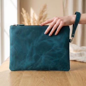 May include: A rectangular teal leather clutch bag featuring a smooth, matte finish with visible natural grain texture. The bag includes a matching teal leather wrist strap attached to a silver-toned metal clasp on the side and a top zipper closure.