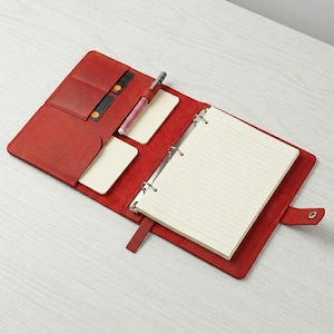 May include: A red leather planner with a metal ring binder mechanism holding lined paper. The interior features multiple card slots, two rectangular pockets, and a pen loop containing a silver and pink pen. A snap button closure is visible.