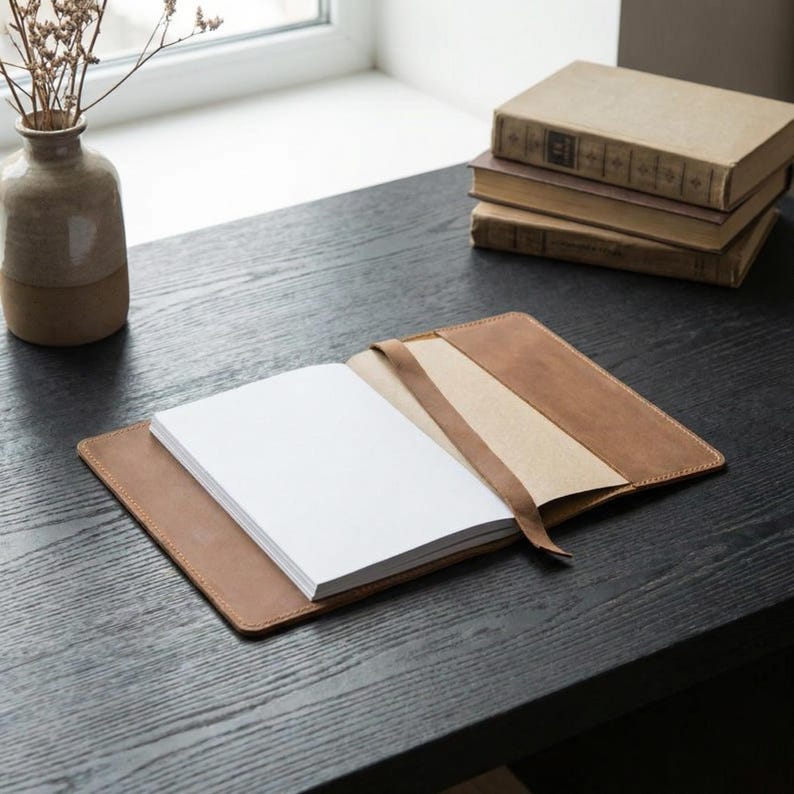 May include: An open, brown leather journal with a stack of white pages and a matching ribbon bookmark. The journal is on a dark wooden surface, with a stack of vintage books and a decorative vase in the background.