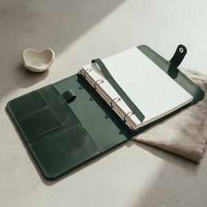 May include: A dark green leather binder featuring a metal ring mechanism, internal pockets, and a snap button closure. The binder is open, revealing a stack of blank white paper pages held in place by a matching green leather strap.