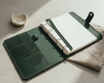 May include: A dark green leather binder featuring a metal ring mechanism, internal pockets, and a snap button closure. The binder is open, revealing a stack of blank white paper pages held in place by a matching green leather strap.