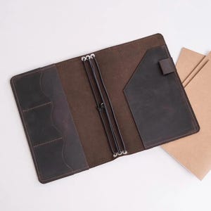 May include: Open, dark brown leather journal cover with multiple pockets and elastic closures. The cover is open, revealing a brown interior and a pocket on the right side. A few blank notebooks are visible on the right.
