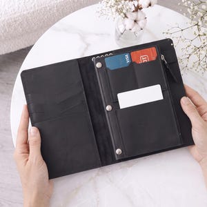 May include: A black leather wallet organizer featuring multiple card slots, a zippered compartment, and a central pocket. The interior includes a blue Visa card, a red card, and a blank white card. The wallet is held open to display its internal structure.