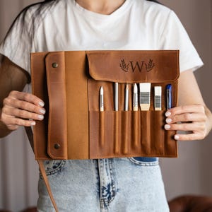 May include: A brown leather artist's roll-up case with a flap, holding various art supplies. The case features a stamped "W" with a leaf design. The case contains paint brushes, pencils, and an eraser. The case is held open by a person.