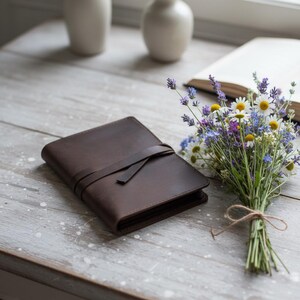 May include: A dark brown leather journal with a strap closure rests on a weathered wooden surface. A bouquet of wildflowers, including daisies and lavender, is tied with twine. Two white vases and an open book are in the background.