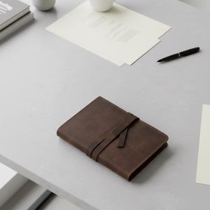 May include: A brown leather journal with a strap closure lies on a gray surface. The journal is rectangular and appears to be closed. A black pen and papers are also on the surface, along with a small stack of books and a white cup.
