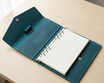 May include: A teal leather binder with a snap closure, featuring an interior pen loop and a pocket on the left side. The binder contains a set of white, unlined pages held in place by a silver-coloured six-ring metal mechanism for organisation.