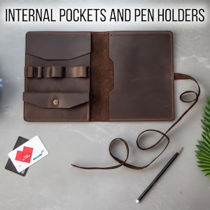May include: Open brown leather folio with the text "INTERNAL POCKETS AND PEN HOLDERS". The folio has internal pockets, pen holders, and a button closure. A pencil and a strap are on the right side. Two cards are on the left.
