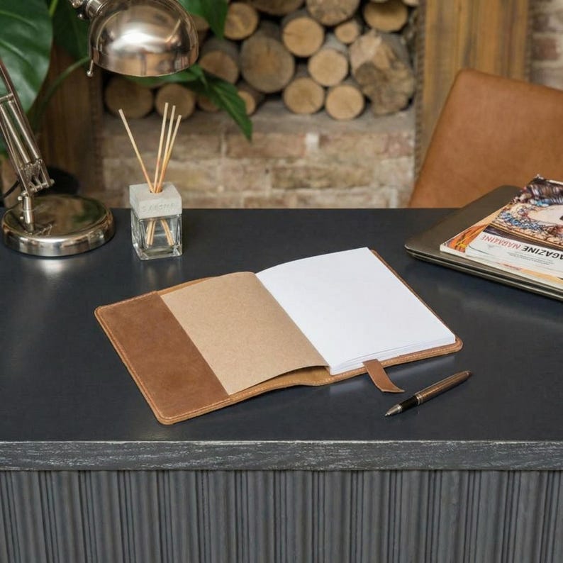 May include: Open brown leather journal with blank white pages and a tan bookmark. A silver pen rests beside the journal on a dark gray desk. A silver desk lamp and a magazine are also visible.