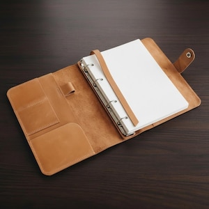 May include: Open, tan leather binder with a ring mechanism holding white paper. The binder has a strap closure and several pockets. The background is a dark brown wooden surface.