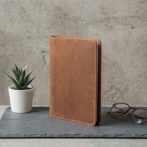 May include: A brown leather journal with a stitched edge, standing upright on a dark gray slate. A small potted succulent and a pair of eyeglasses are also on the slate, against a textured gray background.