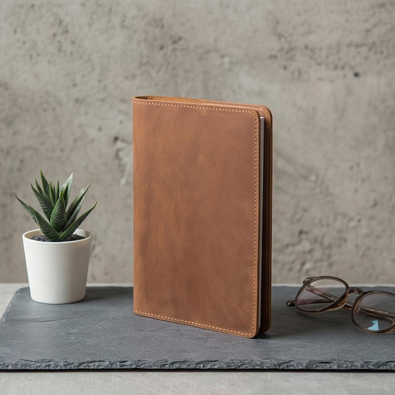 May include: A brown leather journal with a stitched edge, standing upright on a dark gray slate. A small potted succulent and a pair of eyeglasses are also on the slate, against a textured gray background.