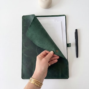 May include: A dark green leather document folder with a snap button closure, containing a stack of white paper. A black pen is positioned to the right of the folder, and a small, round, light-colored ceramic object sits above the folder.