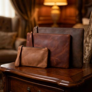 May include: Four leather pouches in varying shades of brown, from light tan to dark brown, are arranged on a wooden surface. Each pouch has a zipper closure, and the smallest one includes a wrist strap. The pouches appear to be made of high-quality leather.