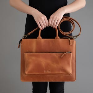 May include: A tan leather tote bag with a top handle and a detachable shoulder strap. The bag features a front pocket with a zip and the initials "D.M." embossed on the front. The bag is held by a person.