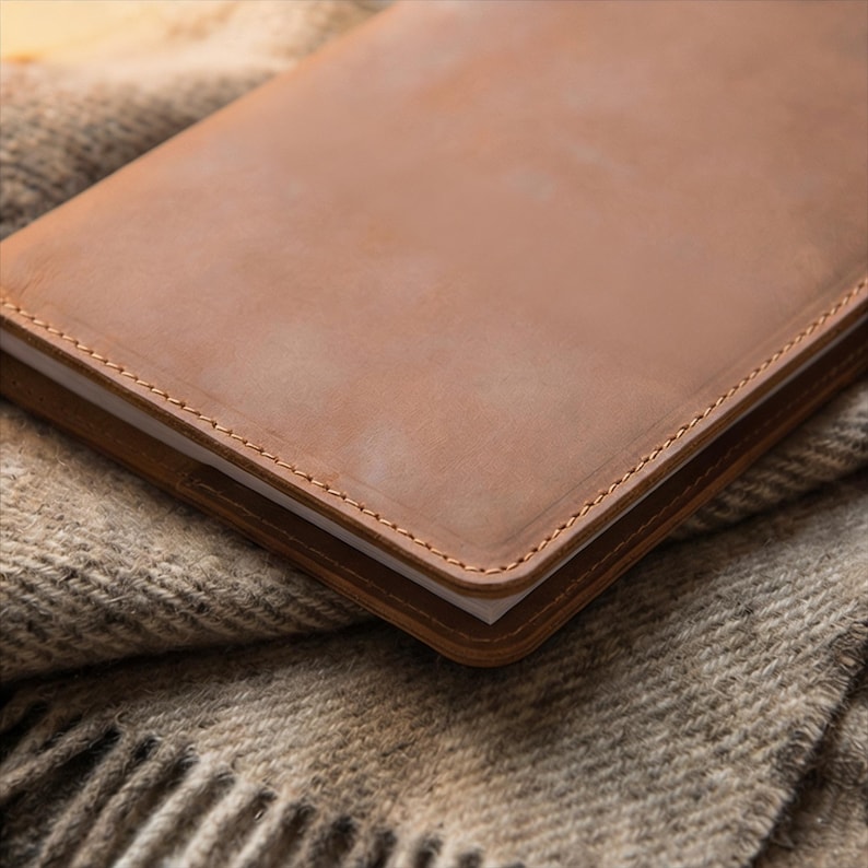 May include: A close-up of a leather-bound notebook with a light brown cover. The notebook is resting on a textured, gray and brown blanket. The edges of the notebook are stitched with a contrasting thread.