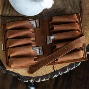 Könnte beinhalten: Ein braunes Lederetui für Mundharmonikas mit mehreren einzelnen Fächern zur Aufbewahrung. Das Etui ist geöffnet und zeigt drei silberne Mundharmonikas in den Taschen sowie einen langen Lederriemen mit Schnallenlöchern für einen sicheren Verschluss.