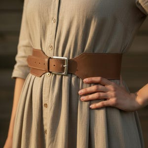 May include: A wide brown leather belt with a silver-colored buckle. The belt is worn over a light brown dress. The belt has a rectangular buckle and is designed to cinch the waist. The belt is approximately 4 inches wide.