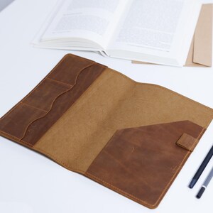 May include: Open leather journal cover in shades of brown. The cover has multiple pockets and a flap closure. An open book and pens are in the background. The journal cover is on a white surface.