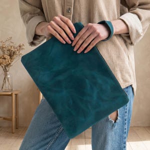 May include: A teal leather clutch bag with a textured finish. The rectangular bag has a zip closure and a matching teal wrist strap. The bag is held against a background of a beige corduroy shirt and blue jeans.