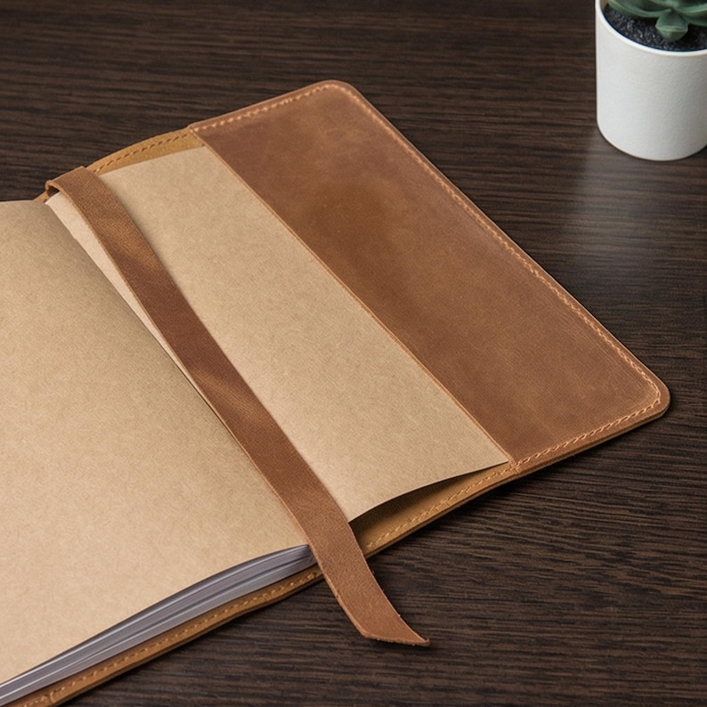 May include: Open brown leather journal with a matching ribbon bookmark. The journal's pages are a light tan color, and the cover has a stitched border. The journal is open, revealing the blank pages inside. A small plant in a white pot is in the background.