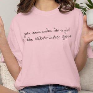 May include: A light pink t-shirt with the handwritten text "you seem calm for a girl in the ticketmaster queue". The shirt is made of a soft material and has short sleeves.