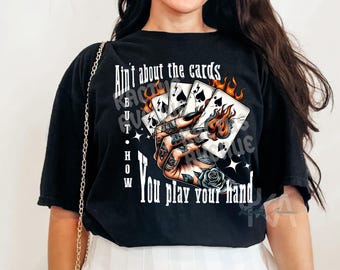 May include: Black t-shirt featuring a graphic of playing cards on fire, held by a hand with tattoos. The text reads "Ain't about the cards but how you play your hand."