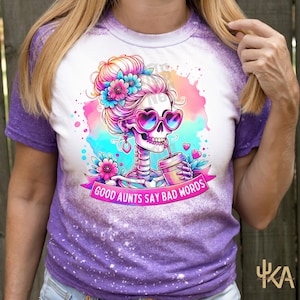 May include: A purple and white t-shirt featuring a colorful illustration of a skeleton with pink heart-shaped sunglasses, flowers, and a coffee cup. The text "GOOD AUNTS SAY BAD WORDS" is on a banner.