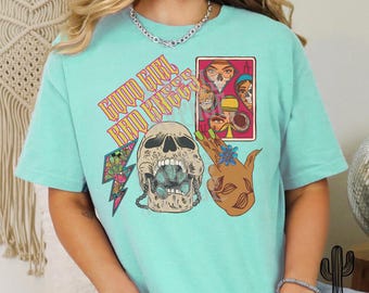 May include: A turquoise blue t-shirt with a graphic print of a skull with a crown, a hand with a flower, and a playing card with a skull and crossbones. The text "Good Girl Bad Kings" is printed on the shirt.