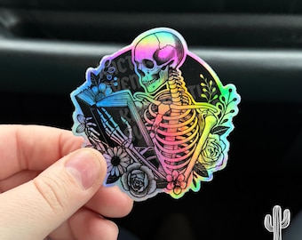 May include: A holographic sticker featuring a skeleton reading a book surrounded by flowers. The skeleton is rainbow colored and the flowers are in shades of pink, purple, and blue.