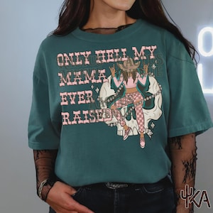 May include: Teal-colored t-shirt with the text "ONLY HELL MY MAMA EVER RAISED" in pink. The graphic features a cowgirl on a skull, wearing a cowboy hat, pink top, and checkered pants.