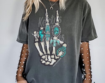 May include: Dark gray t-shirt featuring a skeletal hand with turquoise and silver jewelry. The design includes a turquoise ring, a turquoise and silver pendant, and a turquoise cross. The text "Kaktus Avenue" is printed on the shirt.