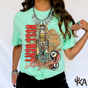 May include: A mint green t-shirt featuring a whiskey-themed graphic. The design includes a whiskey bottle, a pool ball with flames, and the words "Rebel" and "Whiskey Bound" in a western-style font. The shirt has short sleeves.
