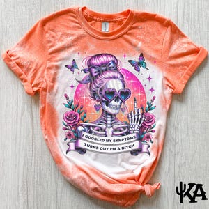 Puede incluir: Camiseta naranja tie-dye con efecto blanqueado. La camiseta presenta un gráfico de un esqueleto con cabello rosado usando gafas en forma de corazón y haciendo el dedo medio. El texto en la camiseta dice "I Googled My Symptoms, Turns Out I'm A Bitch."