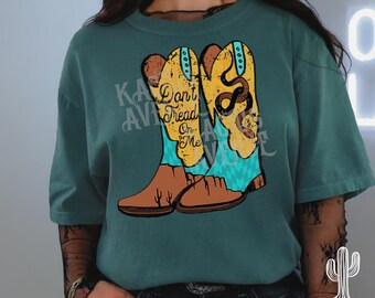 May include: Teal-colored t-shirt featuring a graphic of cowboy boots with a snake and the text "Don't Tread On Me." The boots are yellow and brown with turquoise accents. The shirt has short sleeves.