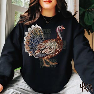 May include: A black long-sleeve sweatshirt featuring a large, detailed graphic of a turkey made of multicolored rhinestones. The turkey design displays shades of brown, gold, blue, and white, with a prominent tail fan and textured appearance.