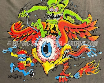 Könnte beinhalten: Ein graues T-Shirt mit einem bunten Grafikdruck eines Monsters, das auf einem geflügelten Auge reitet. Das Monster hat grüne Haut, drei Augen und scharfe Zähne. Das Auge hat eine blaue Iris und rote Adern. Die Flügel sind rot und orange. Das Monster hält einen Schraubenschlüssel und das Auge hält einen Pinsel. Der Text "BACK" ist auf der Unterseite des Shirts gedruckt.