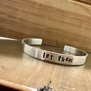 May include: Silver bracelet with the words "let them" engraved on it.