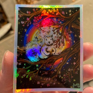 May include: A holographic sticker featuring a white squirrel perched on a tree branch with pink blossoms. A rainbow and full moon are in the background. The sticker has a shiny, iridescent finish and the artist's signature.