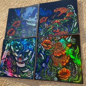 Puede incluir: Cuatro posavasos cuadrados con fondos oscuros, con ilustraciones coloridas. Uno representa una calavera azul con flores y mariposas. Otro muestra una serpiente naranja. Los otros dos presentan diseños florales y esqueléticos.