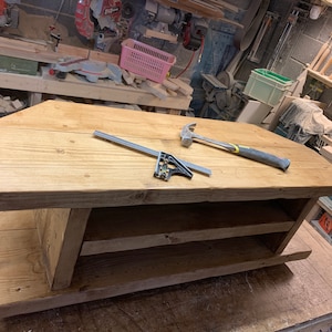 Puede incluir: Una mesa de centro de madera hecha a mano con un acabado natural. La mesa tiene una parte superior rectangular y un estante inferior para almacenamiento. Un martillo y una escuadra est&aacute;n sobre la superficie de la mesa. La mesa est&aacute; en un taller.