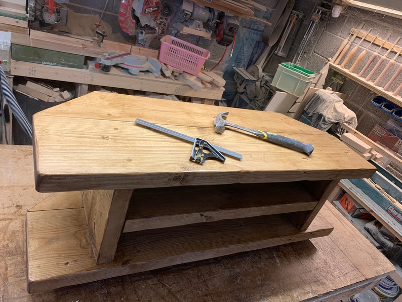 Puede incluir: Una mesa de centro de madera hecha a mano con un acabado natural. La mesa tiene una parte superior rectangular y un estante inferior para almacenamiento. Un martillo y una escuadra est&aacute;n sobre la superficie de la mesa. La mesa est&aacute; en un taller.