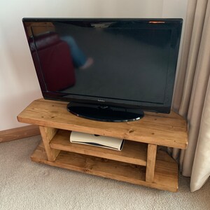 Puede incluir: Un televisor de pantalla plana negro se encuentra sobre un mueble de TV r&uacute;stico de madera marr&oacute;n claro. El mueble tiene dos estantes y est&aacute; hecho de tablones en bruto. El mueble de TV est&aacute; en una esquina de una habitaci&oacute;n con una alfombra de color neutro.