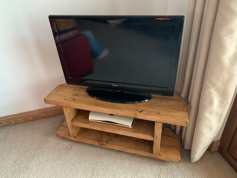 Puede incluir: Un televisor de pantalla plana negro se encuentra sobre un mueble de TV r&uacute;stico de madera marr&oacute;n claro. El mueble tiene dos estantes y est&aacute; hecho de tablones en bruto. El mueble de TV est&aacute; en una esquina de una habitaci&oacute;n con una alfombra de color neutro.